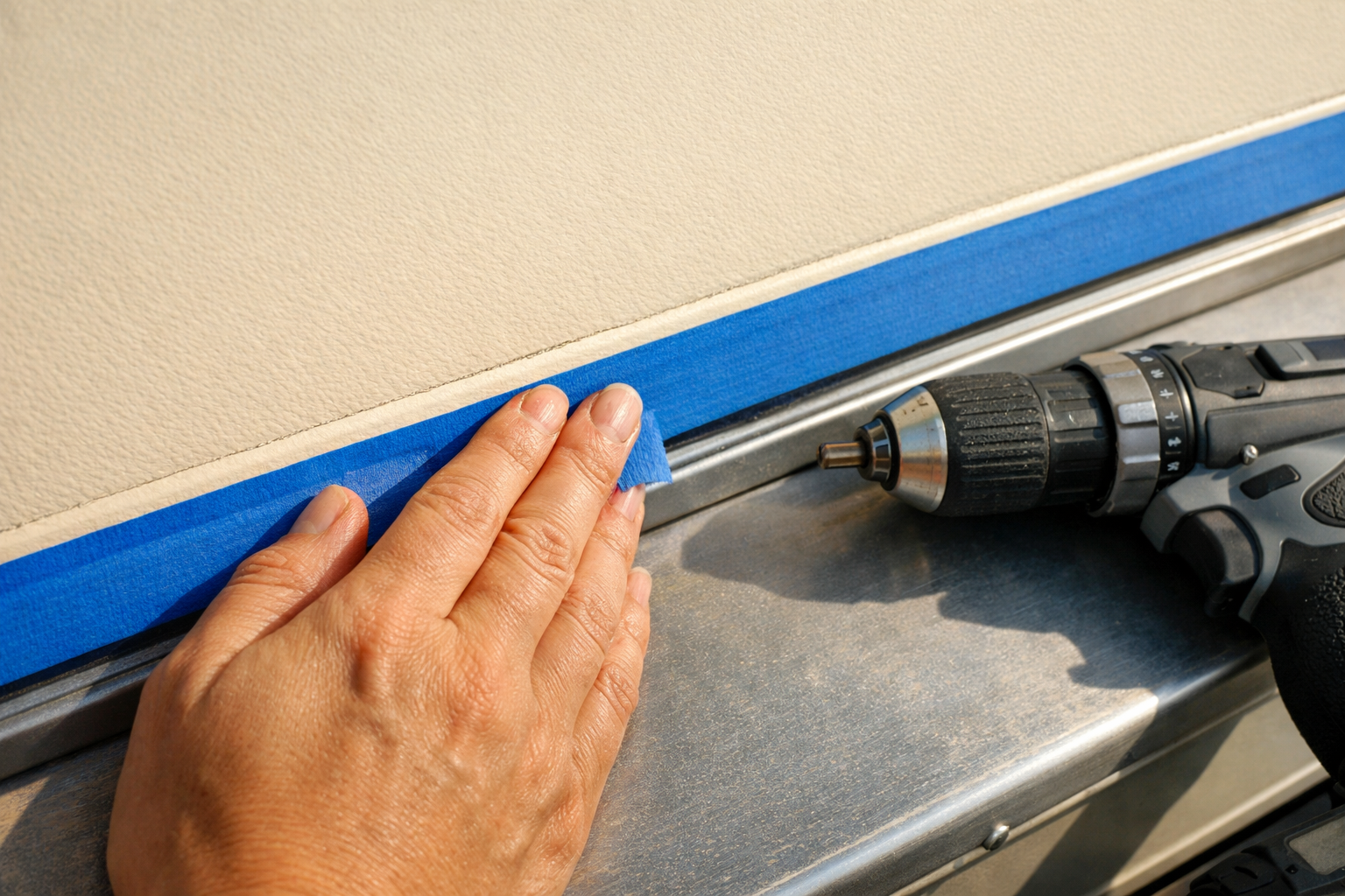 A person's hands actively pressing a strip of blue painter's tape along the edge of a cream-colored RV door panel, fingers smoothing down the tape in a single fluid motion, a cordless drill resting nearby on the metal door surface, natural afternoon light casting soft shadows across the textured door finish, shot from a close overhead angle freezing the mid-press moment, authentic candid style like a DIY enthusiast documenting their own project.